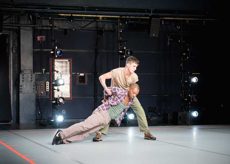 A white dancer holds a black dancer by his shoulders. The black dancer, Kyle Abraham's focus is at the floor. His hands are behind his back.